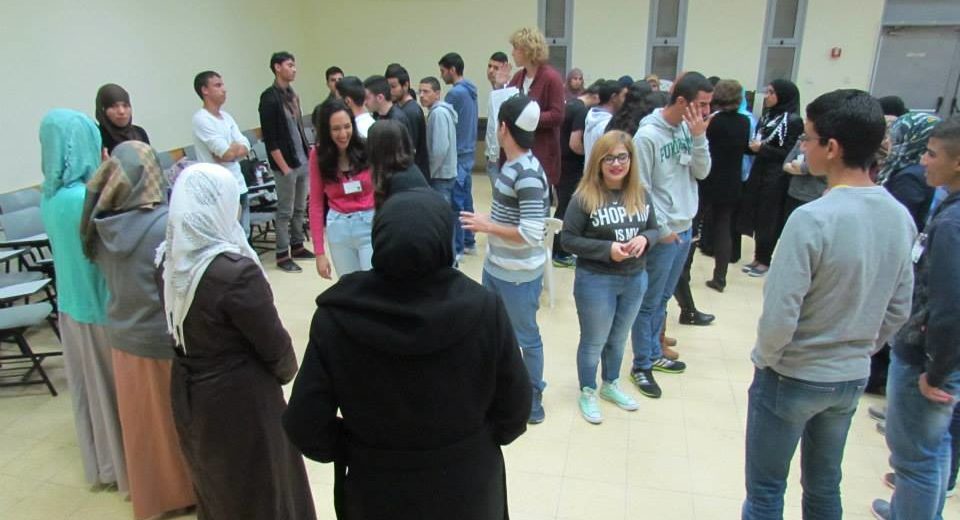 Sharón stands on a chair in the center of a circle of students, leading an introductory game at a Young Negotiators Training seminar with Arab and Jewish Israeli high school students.